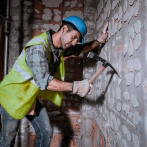 A worker with a helmet on his head hits a wall with a pickaxe in a building under construction.